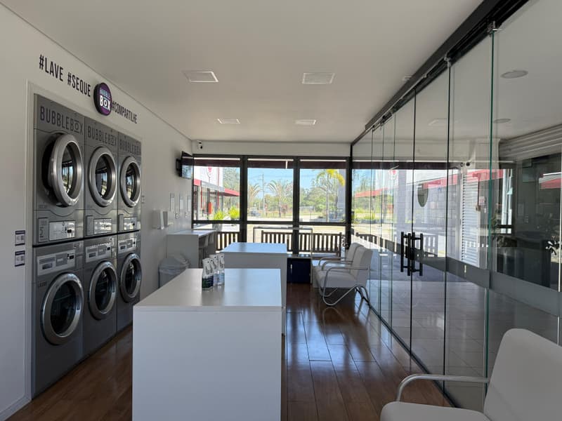 Salão da lavanderia visto do fundo, com a parede de máquinas à esquerda, a mesa de dobrar roupa no centro e as poltronas junto à vitrine que dá para o estacionamento