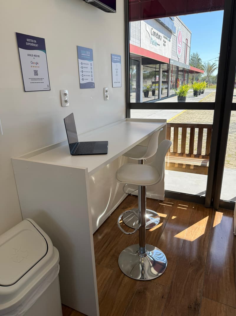 Bancada alta junto à parede com um notebook aberto, uma banqueta e tomadas ao alcance da mão, iluminada por luz natural