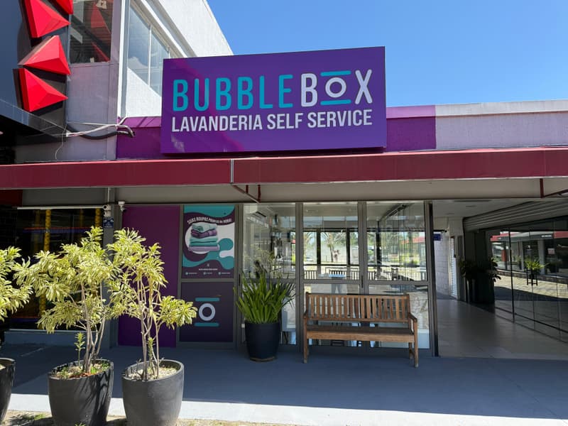Fachada da Bubble Box no Pátio GV, com o letreiro roxo acima da entrada de vidro, um banco de madeira e vasos de plantas na calçada