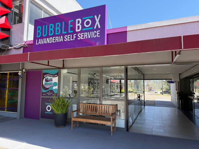 Fachada da Bubble Box vista de lado, com o letreiro roxo sobre a entrada de vidro, um banco de madeira e a calçada coberta do Pátio GV seguindo à direita